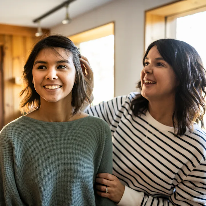 Two smiling women stand in a sunlit room near windows; one adjusts the other's hair while they chat,...