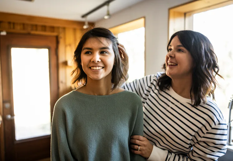 Two smiling women stand in a sunlit room near windows; one adjusts the other's hair while they chat,...