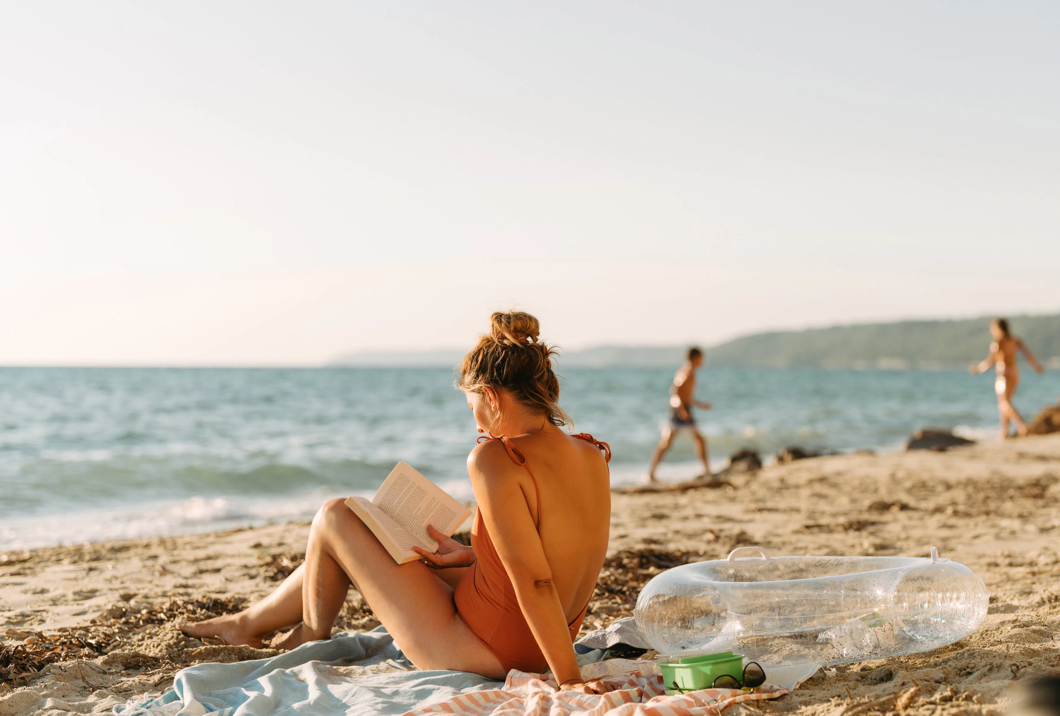 Young woman in an orange swimsuit sits on a towel reading a book on a sunlit sandy beach, with an in...