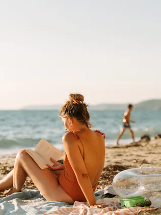Young woman in an orange swimsuit sits on a towel reading a book on a sunlit sandy beach, with an in...