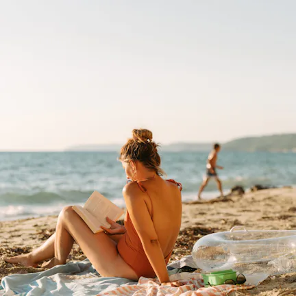 Young woman in an orange swimsuit sits on a towel reading a book on a sunlit sandy beach, with an in...