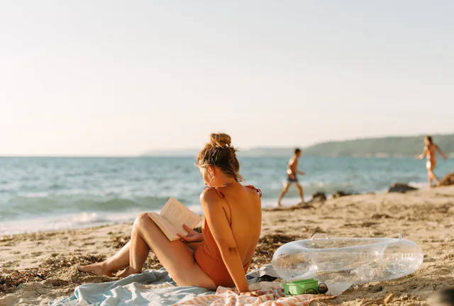 Young woman in an orange swimsuit sits on a towel reading a book on a sunlit sandy beach, with an in...