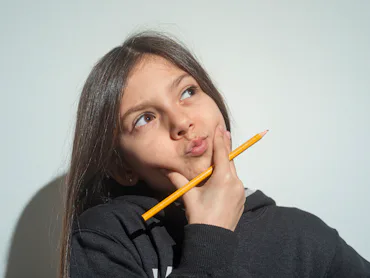 Young girl with long brown hair in a black hoodie looks up thoughtfully, holding a yellow pencil aga...