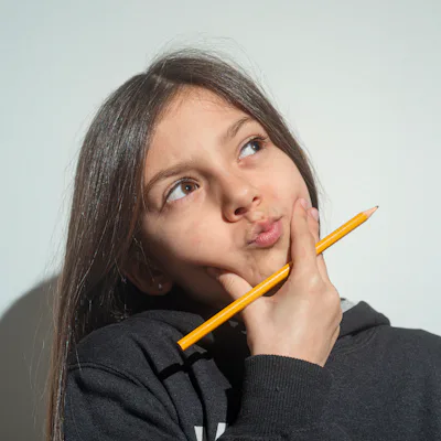 Young girl with long brown hair in a black hoodie looks up thoughtfully, holding a yellow pencil against her cheek and pursing her lips.