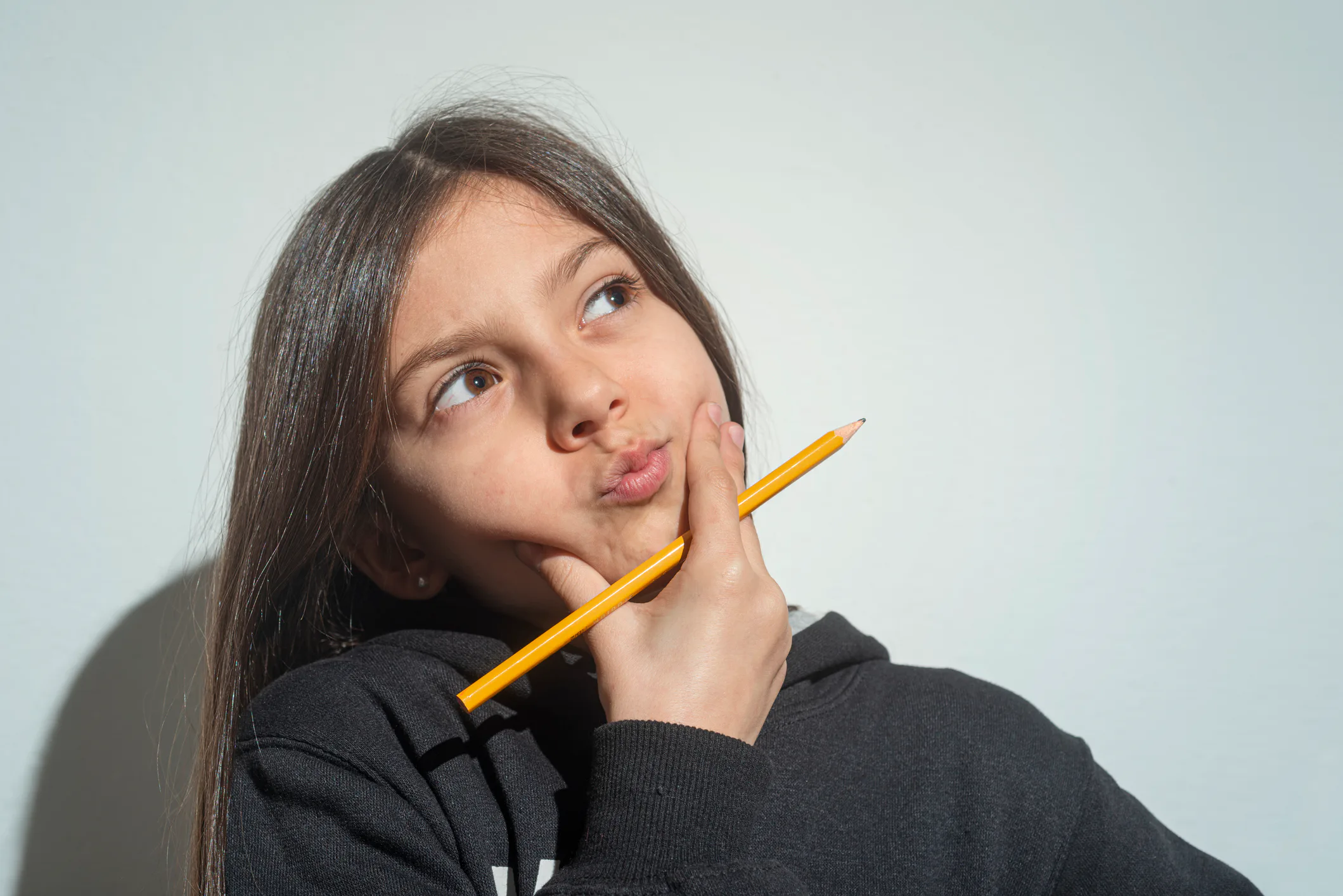 Young girl in a dark hoodie holding a pencil to her chin, looking upward with a thoughtful, pursed e...