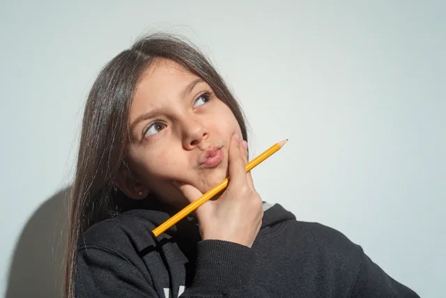 Young girl in a dark hoodie holding a pencil to her chin, looking upward with a thoughtful, pursed e...