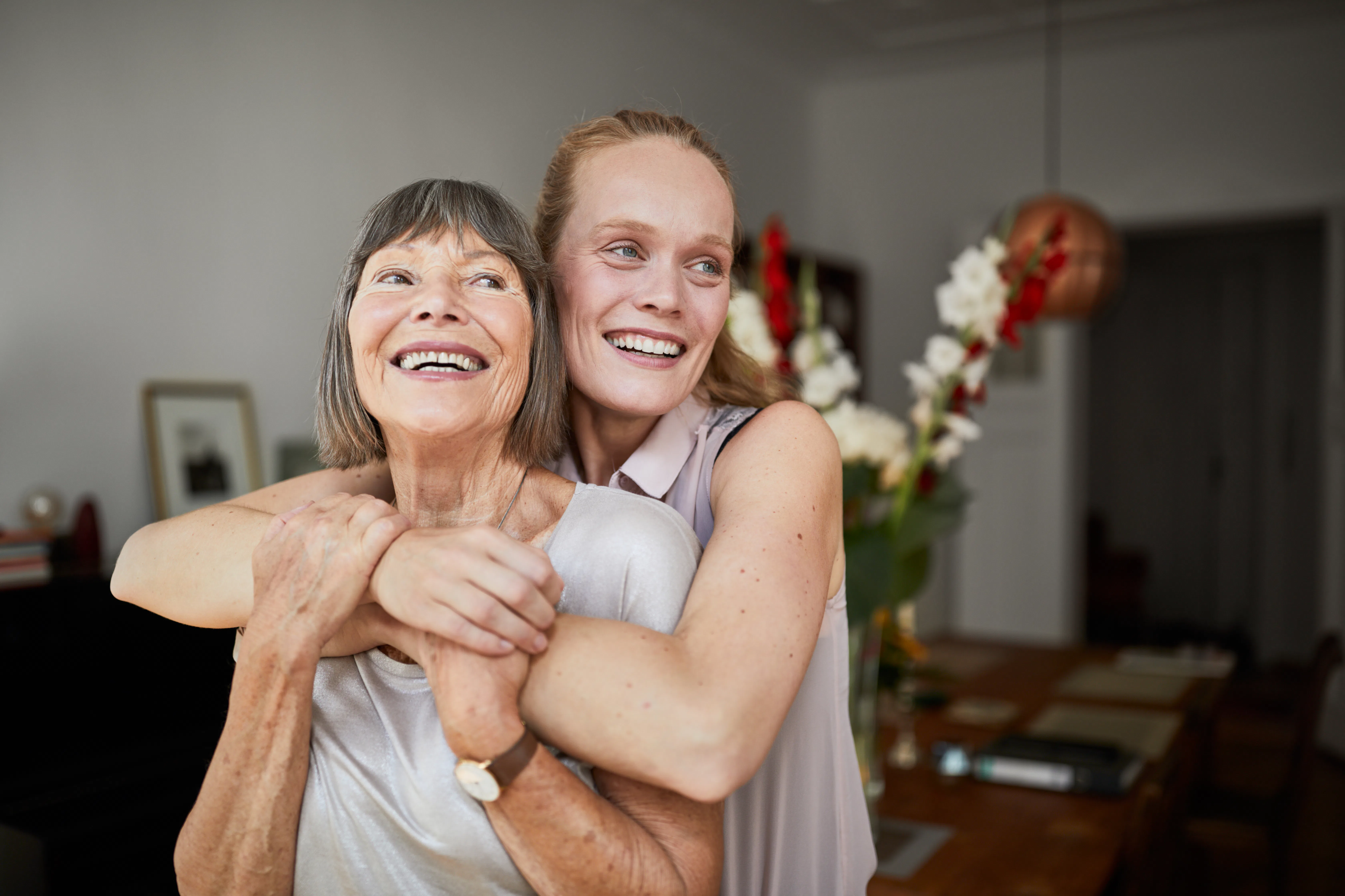 Older woman smiling as a younger woman hugs her from behind in a warmly lit dining room, flowers and...