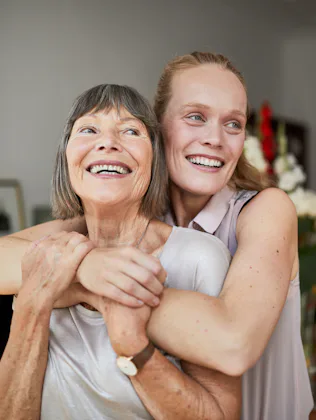 Older woman smiling as a younger woman hugs her from behind in a warmly lit dining room, flowers and...