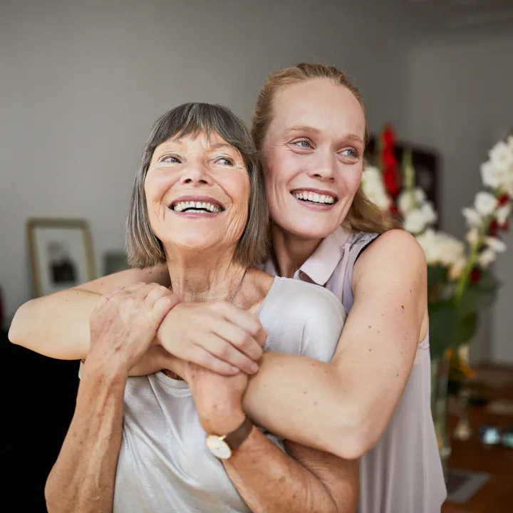Older woman smiling as a younger woman hugs her from behind in a warmly lit dining room, flowers and...