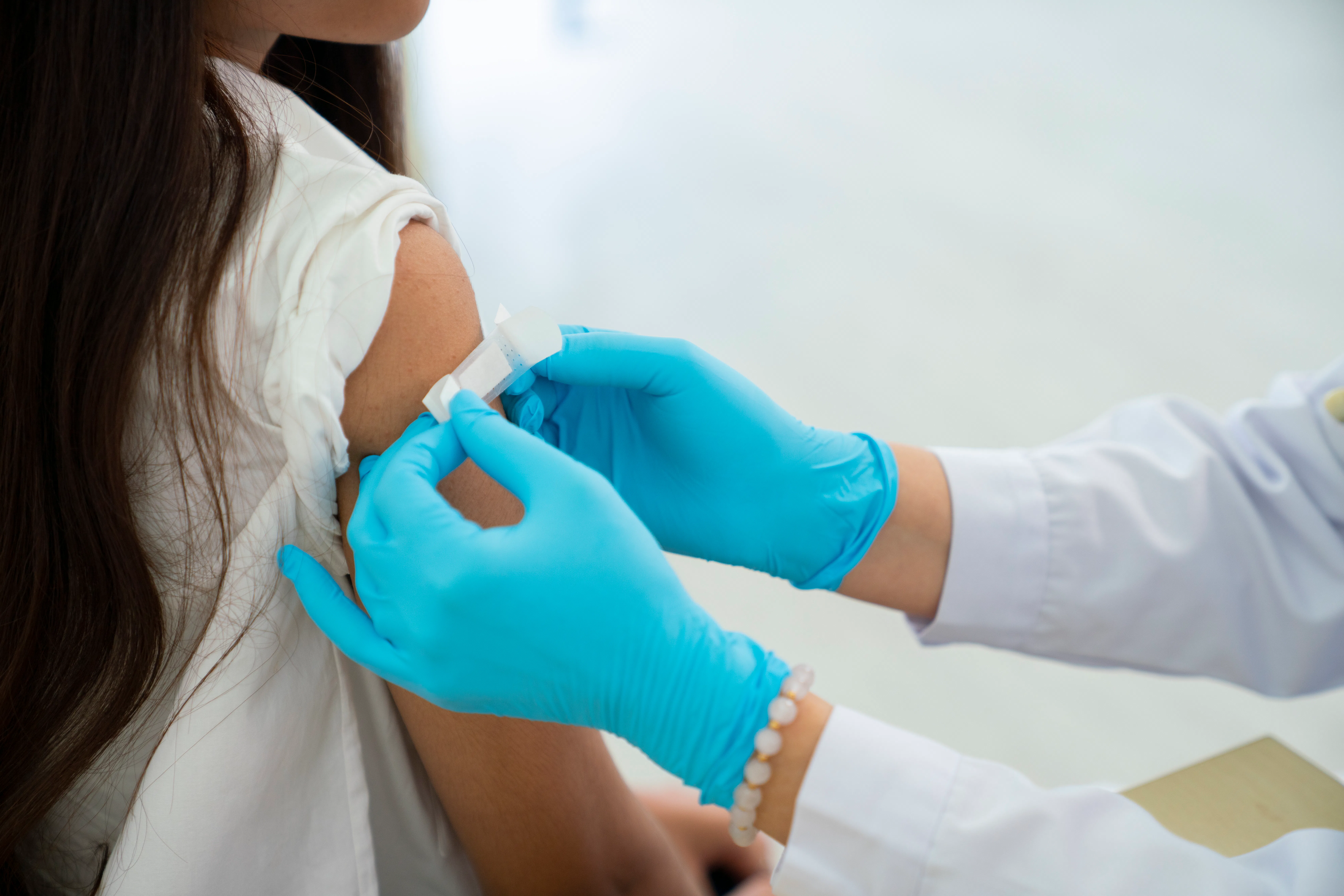 Healthcare worker in blue gloves preparing a bandage on a young person's upper arm after a vaccine o...