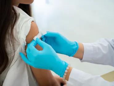 Healthcare worker in blue gloves preparing a bandage on a young person's upper arm after a vaccine o...