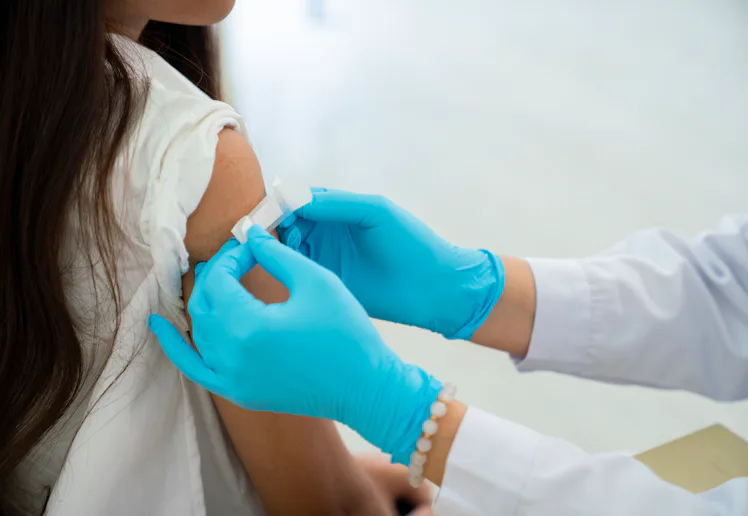Healthcare worker in blue gloves preparing a bandage on a young person's upper arm after a vaccine o...
