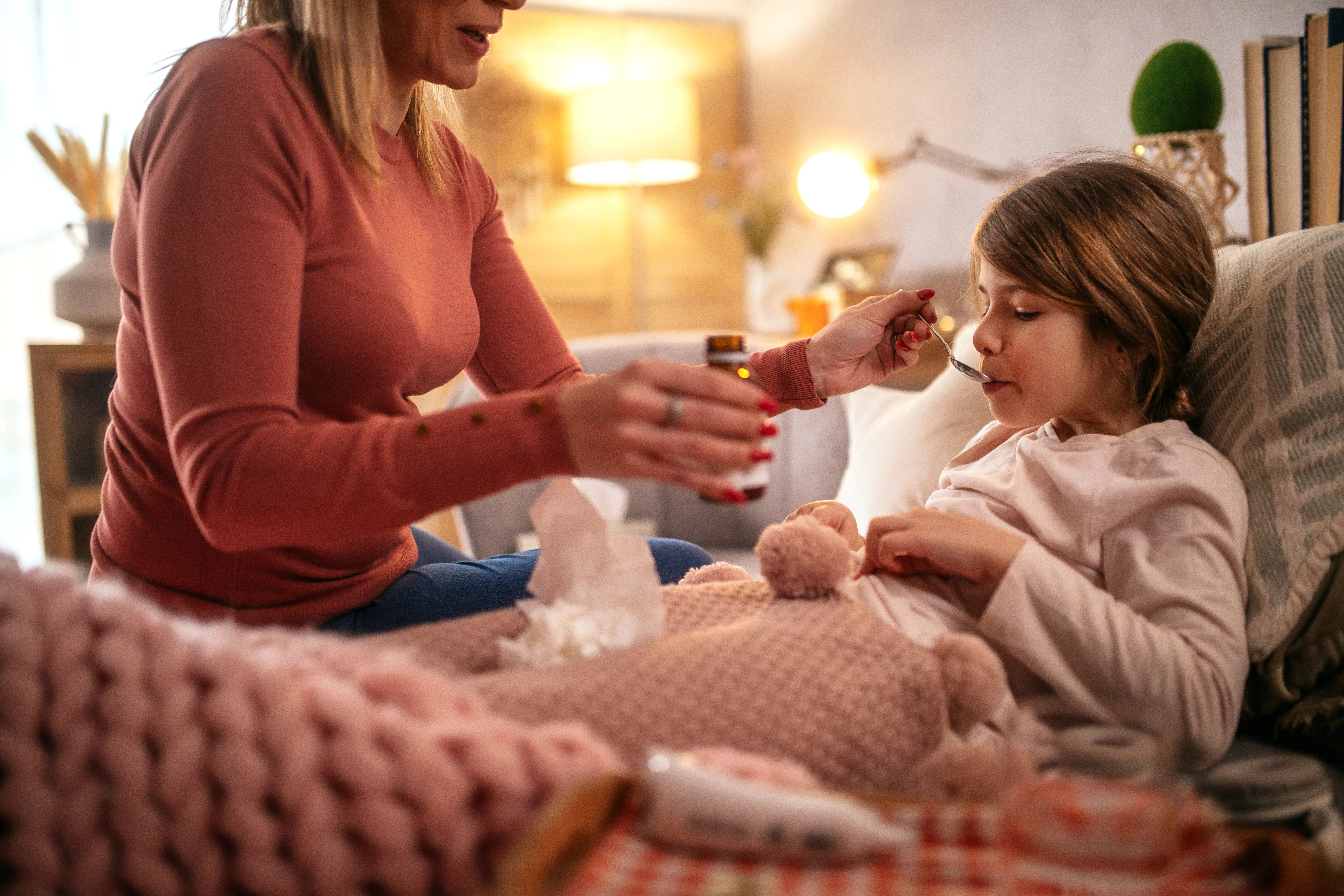 Adult woman, partially seen, feeds a sick child medicine with a spoon as the child sits wrapped in a...