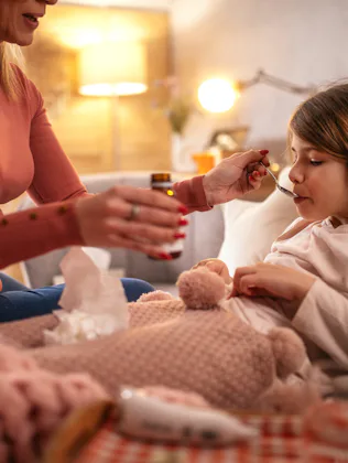 Adult woman, partially seen, feeds a sick child medicine with a spoon as the child sits wrapped in a...