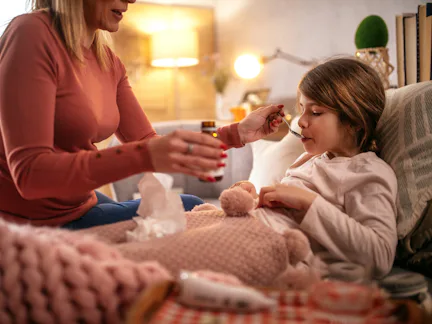 Adult woman, partially seen, feeds a sick child medicine with a spoon as the child sits wrapped in a...