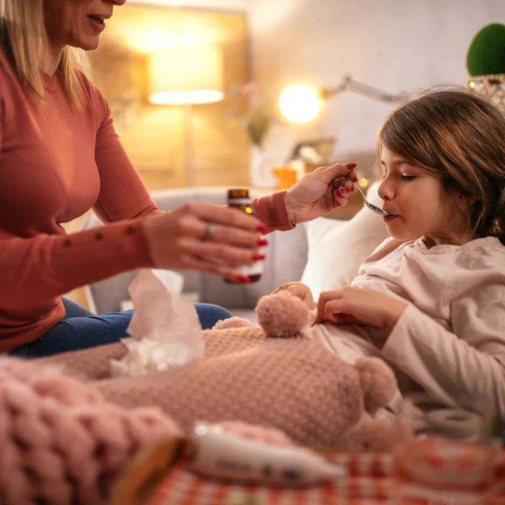 Adult woman, partially seen, feeds a sick child medicine with a spoon as the child sits wrapped in a...