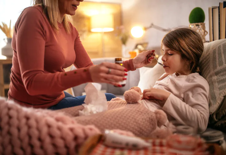 Adult woman, partially seen, feeds a sick child medicine with a spoon as the child sits wrapped in a...