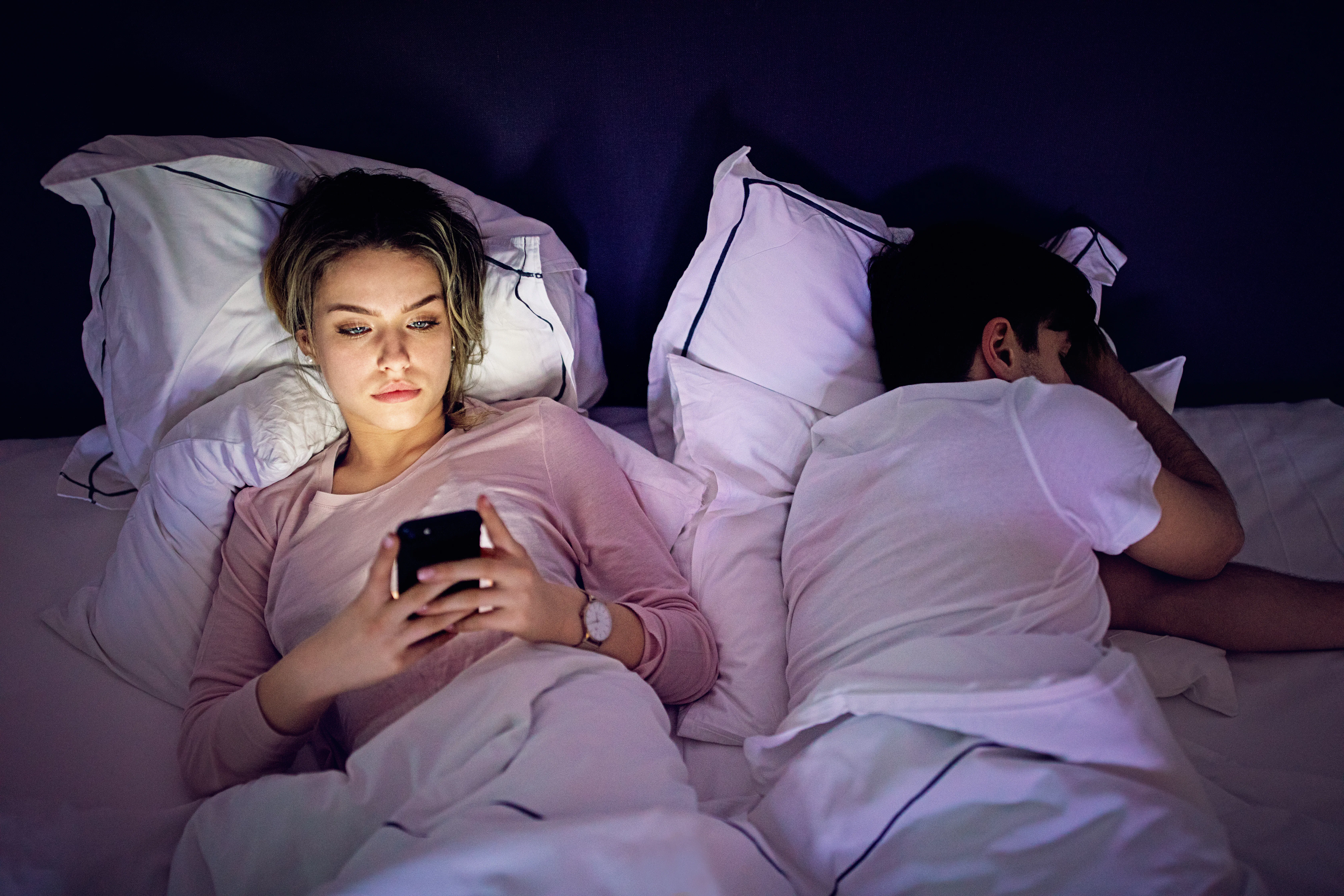 Woman lying awake in bed using a phone, light from the screen on her face; partner sleeps beside her...