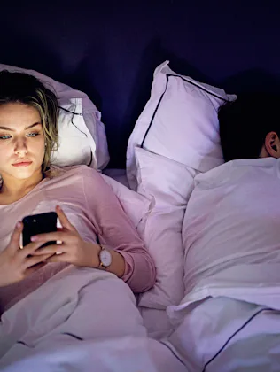 Woman lying awake in bed using a phone, light from the screen on her face; partner sleeps beside her...