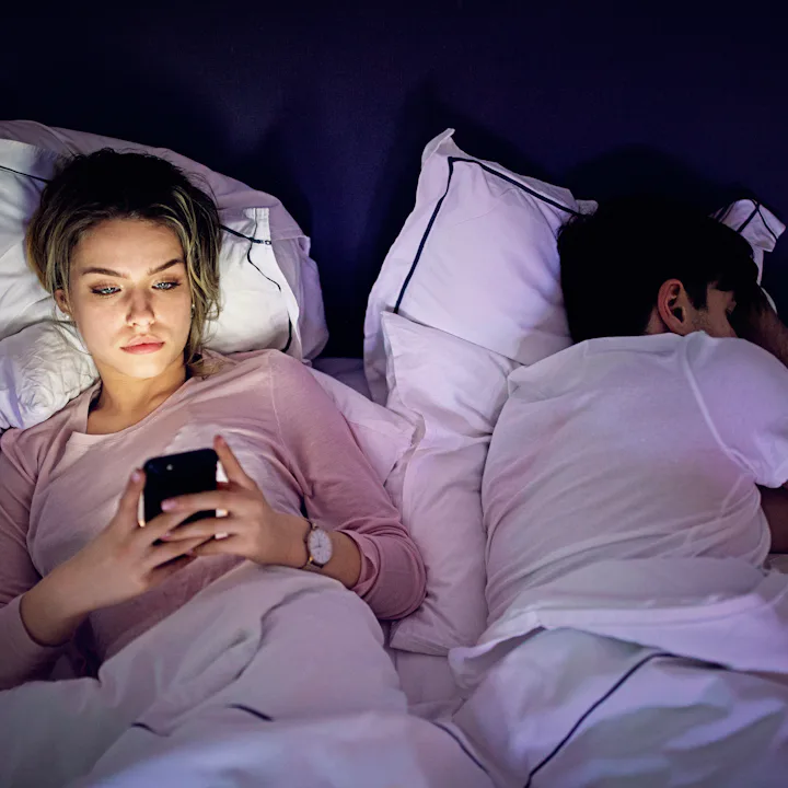 Woman lying awake in bed using a phone, light from the screen on her face; partner sleeps beside her...
