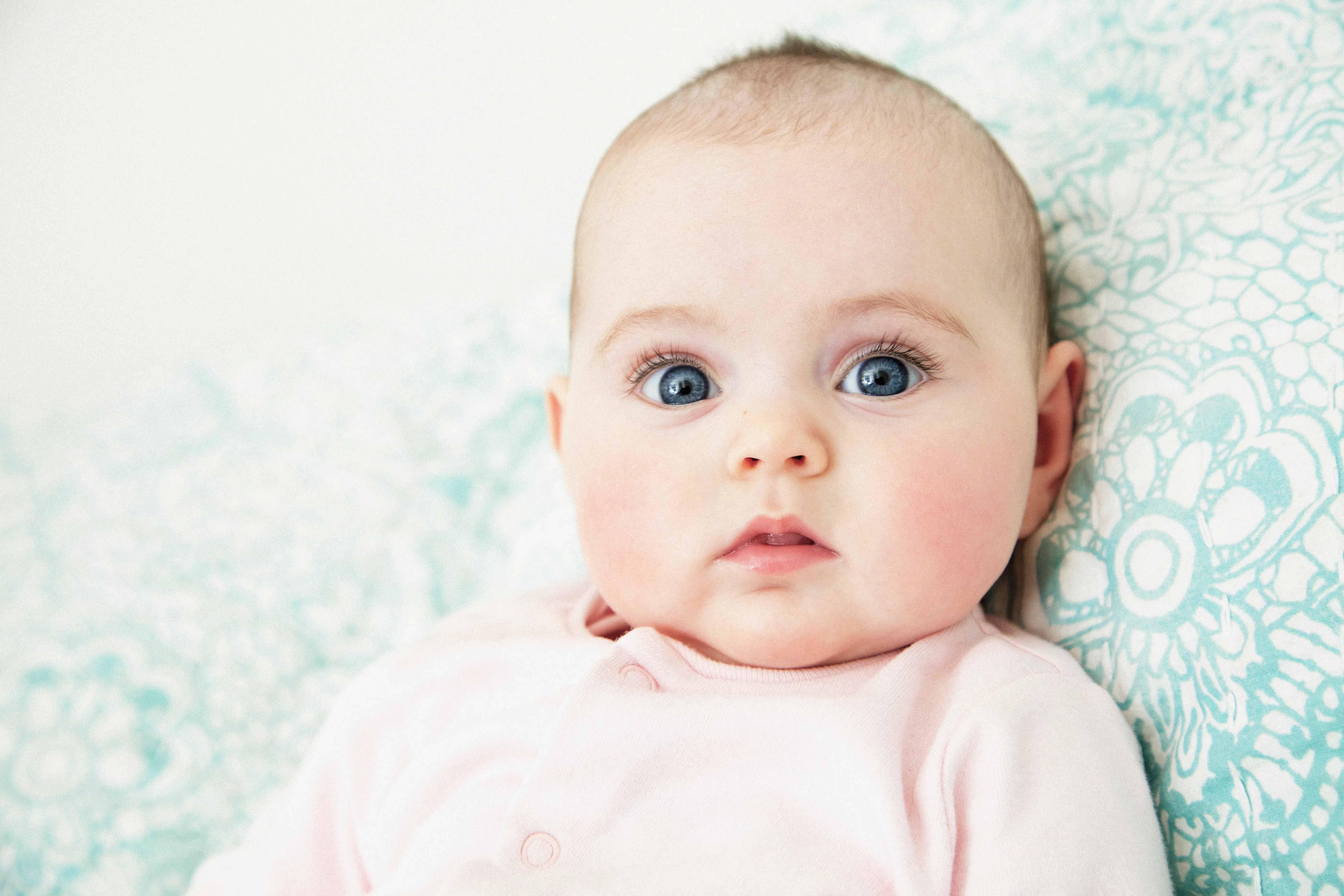 Close-up of a wide-eyed infant with blue eyes and rosy cheeks lying on a patterned teal cushion, wearing a pale pink onesie and looking at the camera.