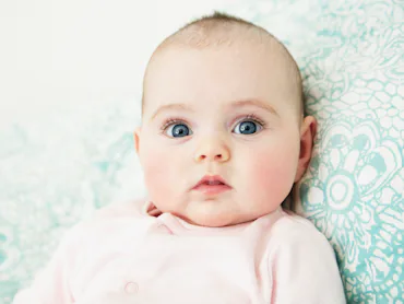Close-up of a wide-eyed infant with blue eyes and rosy cheeks lying on a patterned teal cushion, wea...