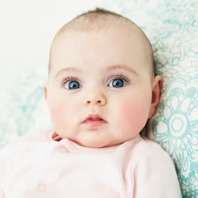 Close-up of a wide-eyed infant with blue eyes and rosy cheeks lying on a patterned teal cushion, wearing a pale pink onesie and looking at the camera.