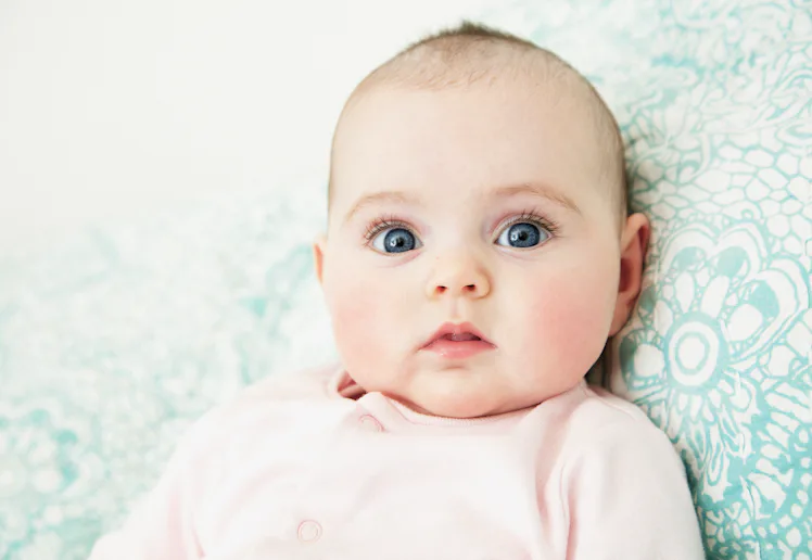 Close-up of a wide-eyed infant with blue eyes and rosy cheeks lying on a patterned teal cushion, wea...
