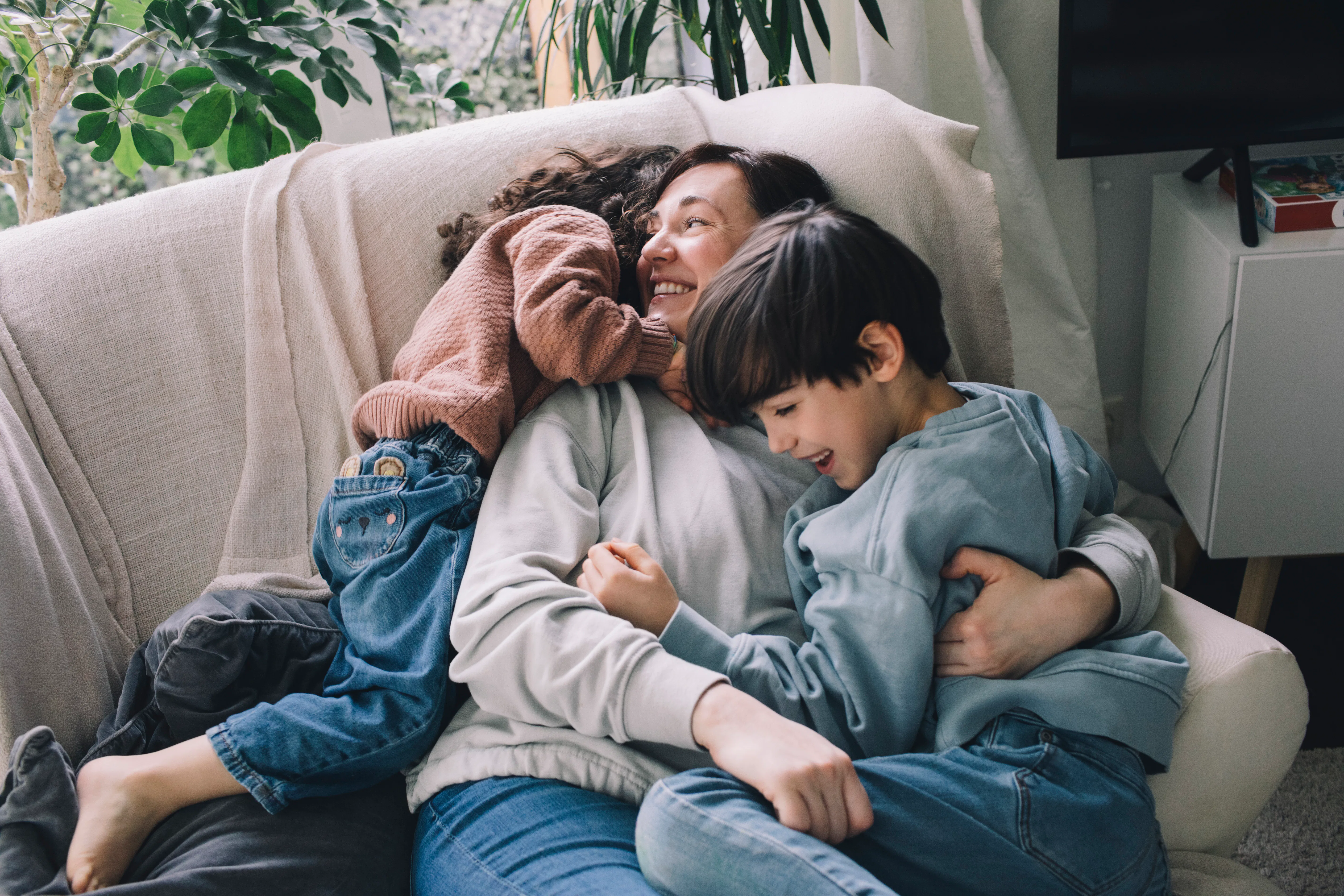 Parent reclining on a light couch, smiling while hugging two young children who cuddle closely; cozy...