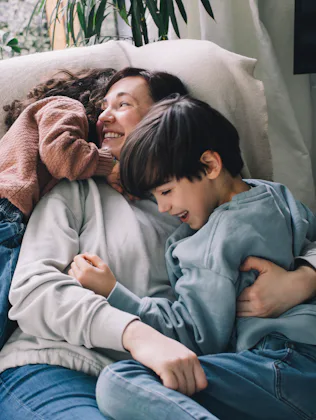 Parent reclining on a light couch, smiling while hugging two young children who cuddle closely; cozy...