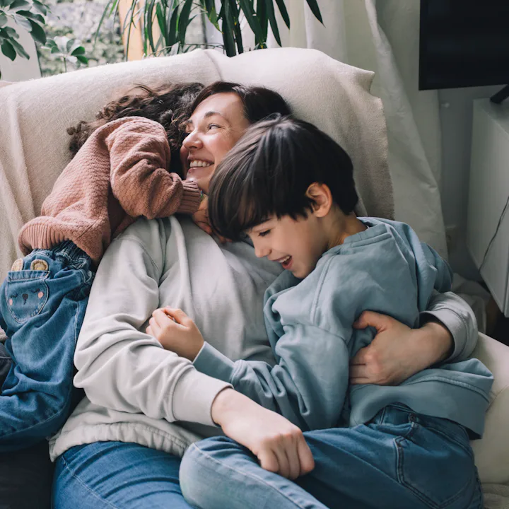 Parent reclining on a light couch, smiling while hugging two young children who cuddle closely; cozy...