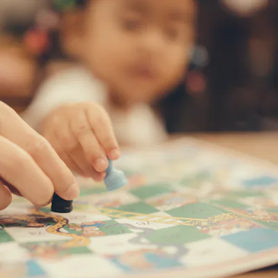 Close-up of an adult and a child moving game pieces on a colorful board game, hands in focus while the child's blurred face watches in background.