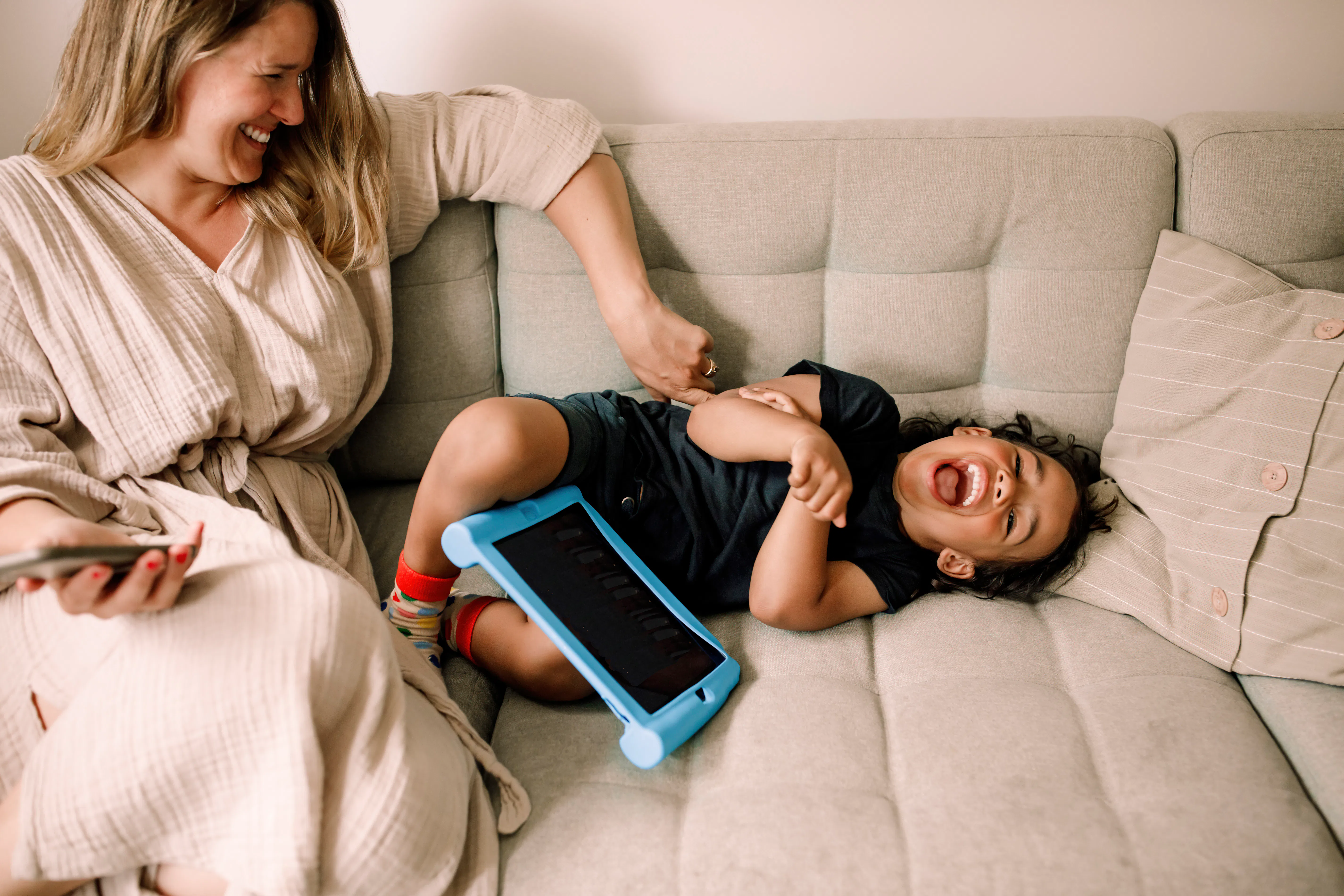 Smiling woman sits on a light gray sofa tickling a laughing toddler lying beside her, a blue tablet ...