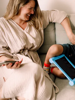 Smiling woman sits on a light gray sofa tickling a laughing toddler lying beside her, a blue tablet ...