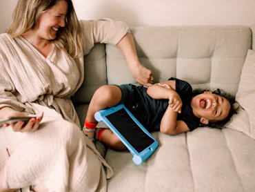 Smiling woman sits on a light gray sofa tickling a laughing toddler lying beside her, a blue tablet ...