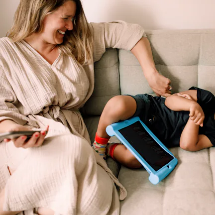 Smiling woman sits on a light gray sofa tickling a laughing toddler lying beside her, a blue tablet ...