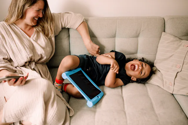 Smiling woman sits on a light gray sofa tickling a laughing toddler lying beside her, a blue tablet ...