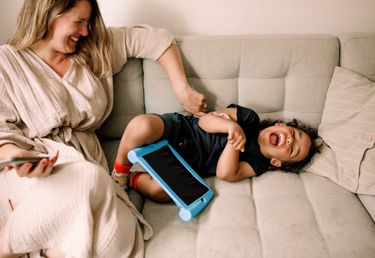 Smiling woman sits on a light gray sofa tickling a laughing toddler lying beside her, a blue tablet ...