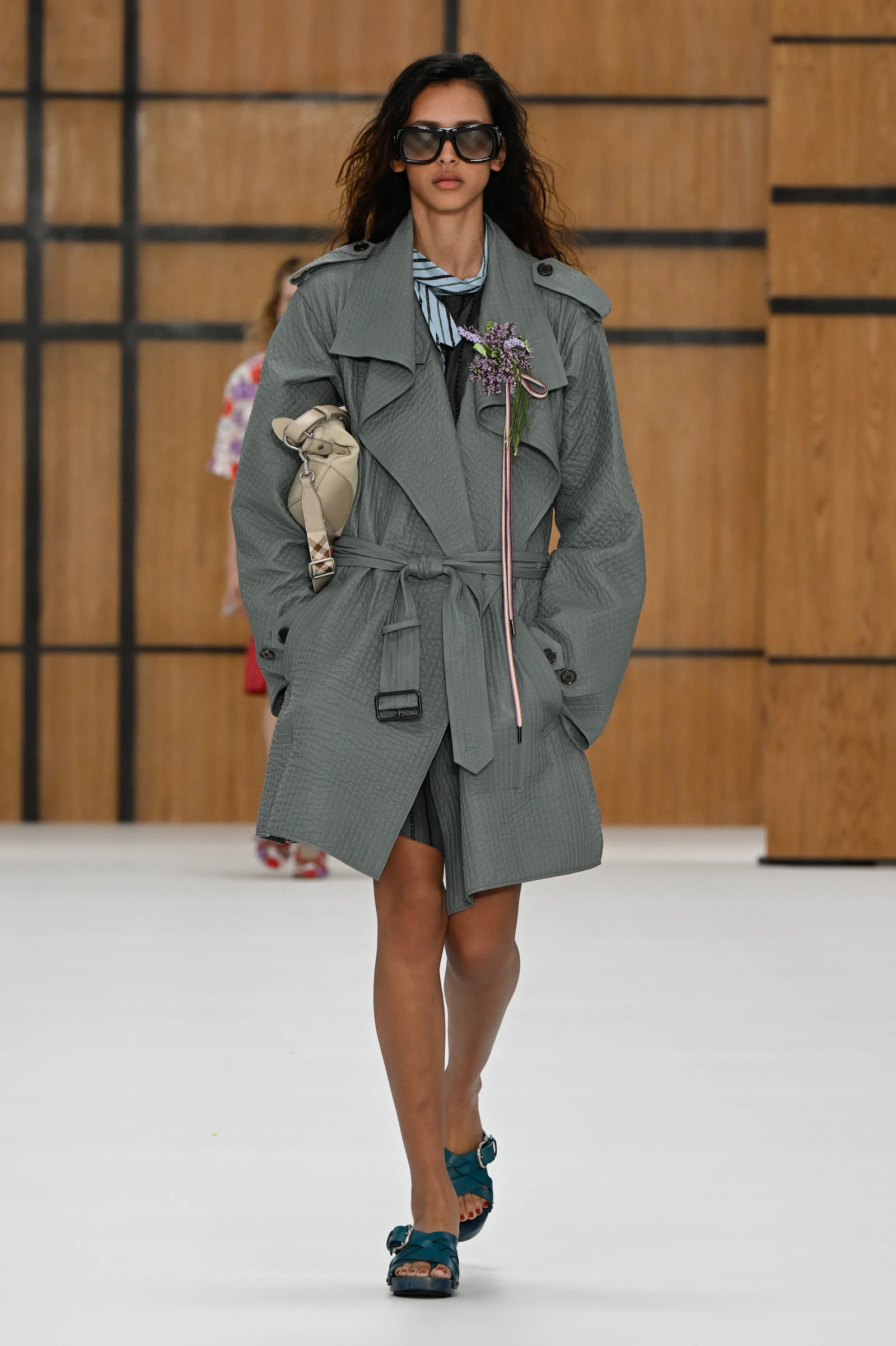 Model in a grey belted Burberry trench styled with sunglasses, teal shoes and a long flower