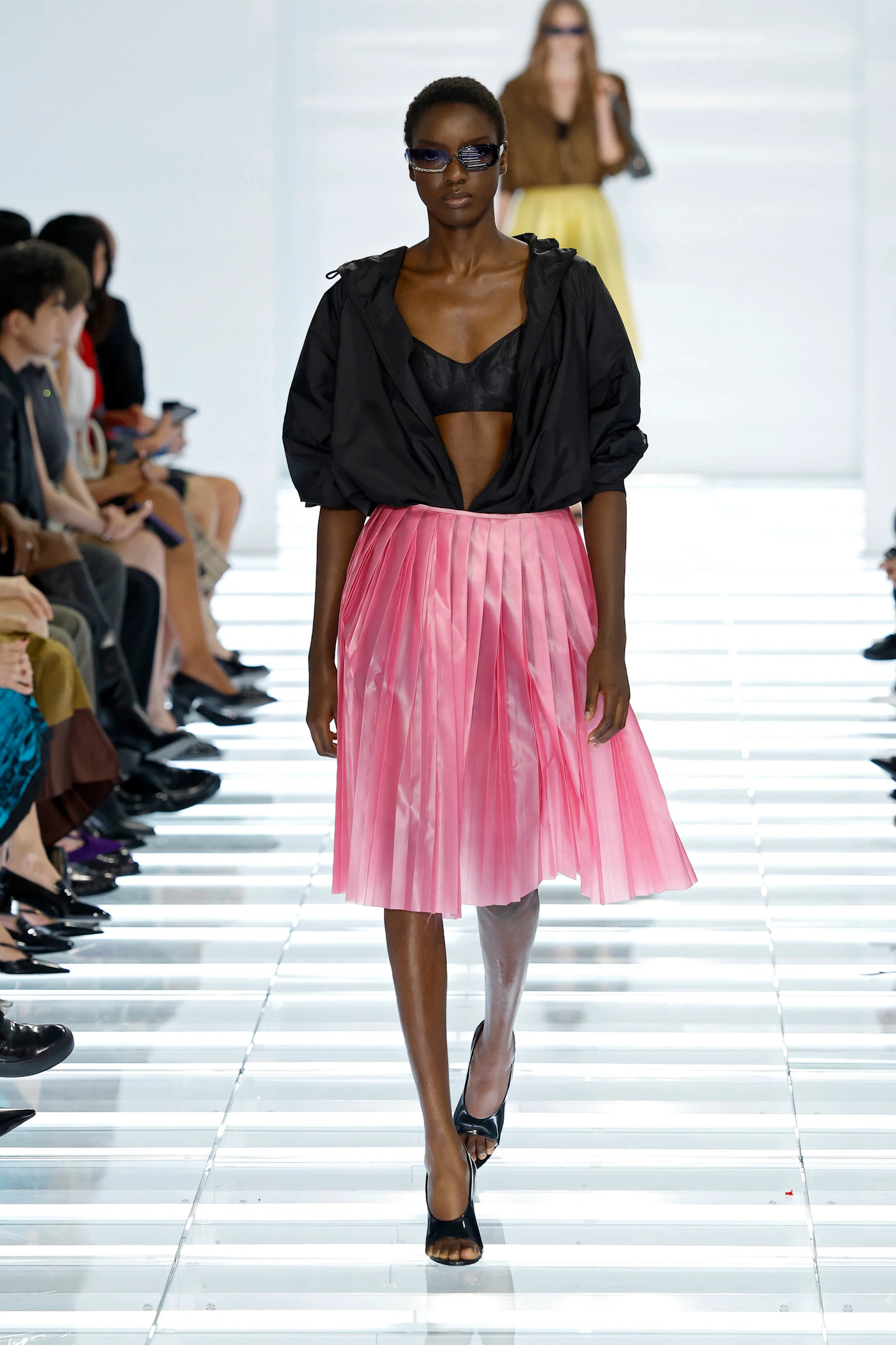 Model in a sheer black Prada blouse and bright pink pleated skirt