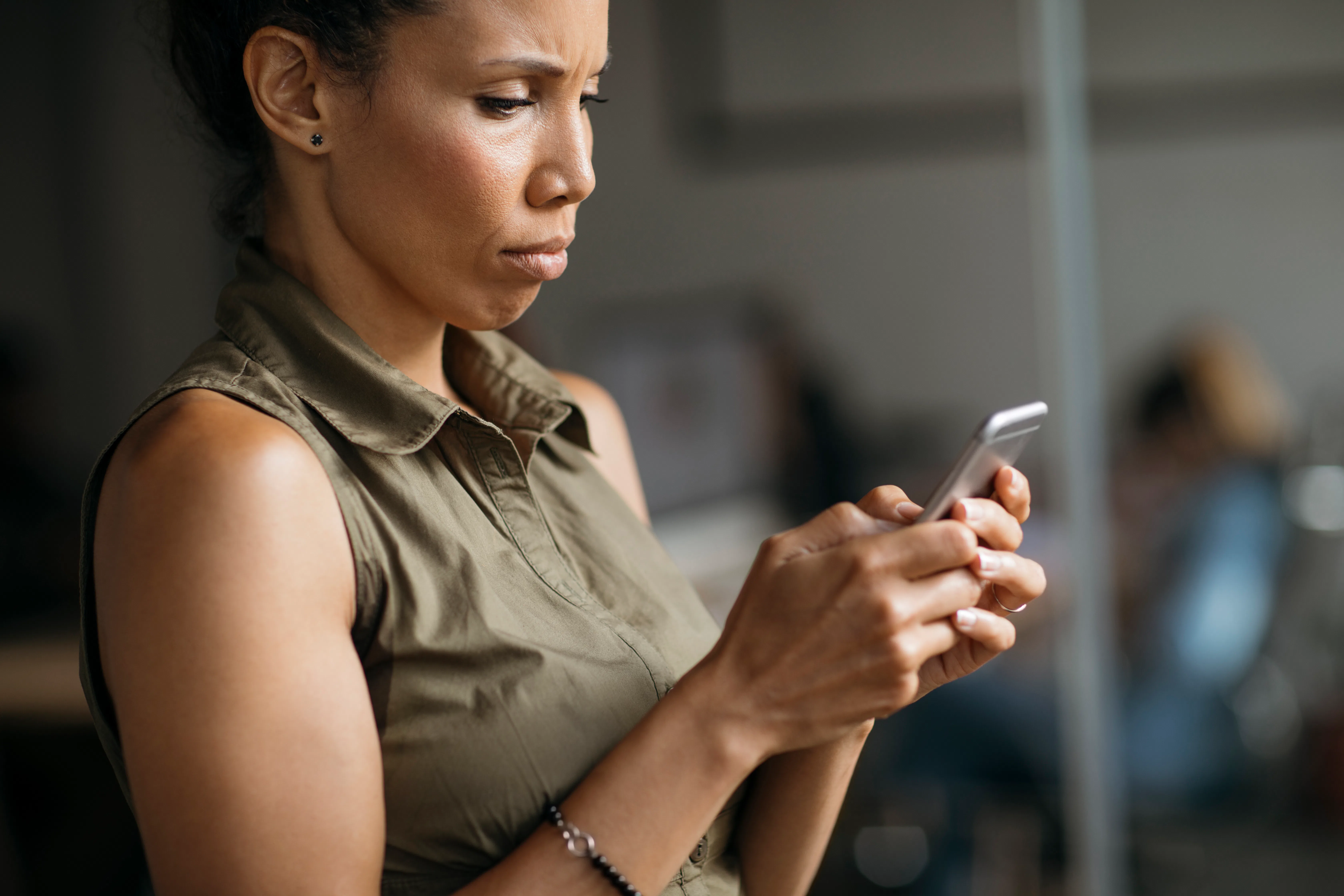 Woman in a sleeveless olive top sitting indoors, focused expression as she holds and types on a smar...