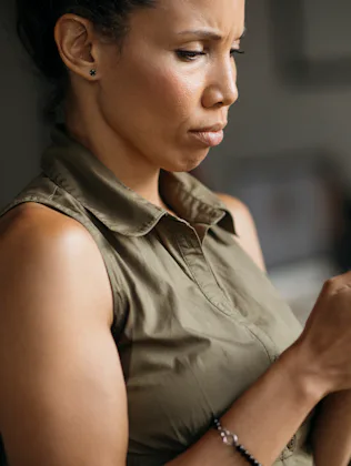 Woman in a sleeveless olive top sitting indoors, focused expression as she holds and types on a smar...