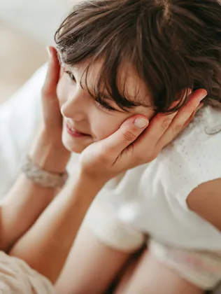 Mother gently cups smiling young girl's face, leaning close in an intimate, warm moment on a bed wit...