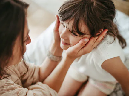Mother gently cups smiling young girl's face, leaning close in an intimate, warm moment on a bed wit...