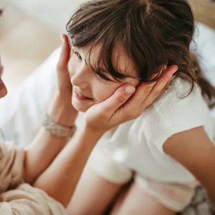 Mother gently cups smiling young girl's face, leaning close in an intimate, warm moment on a bed wit...