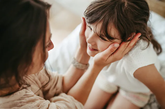 Mother gently cups smiling young girl's face, leaning close in an intimate, warm moment on a bed wit...