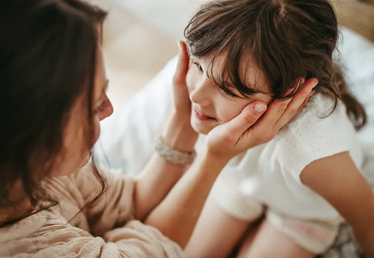 Mother gently cups smiling young girl's face, leaning close in an intimate, warm moment on a bed wit...