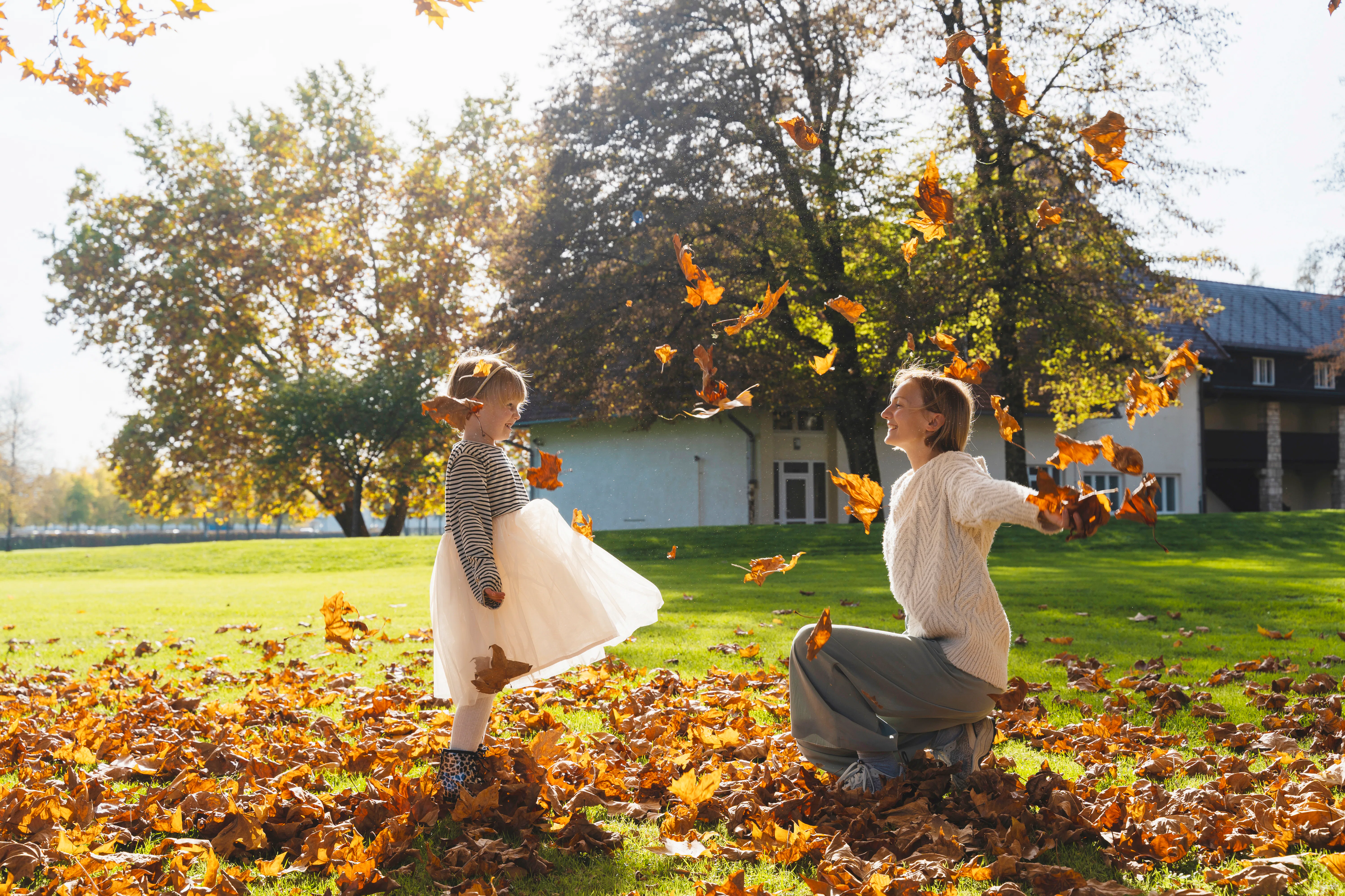 Adult and child playfully tossing autumn leaves in a sunlit yard, surrounded by fallen leaves, trees...
