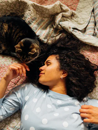 Woman with curly hair lying on a quilted bed, reaching toward a tabby cat curled beside her, both fa...