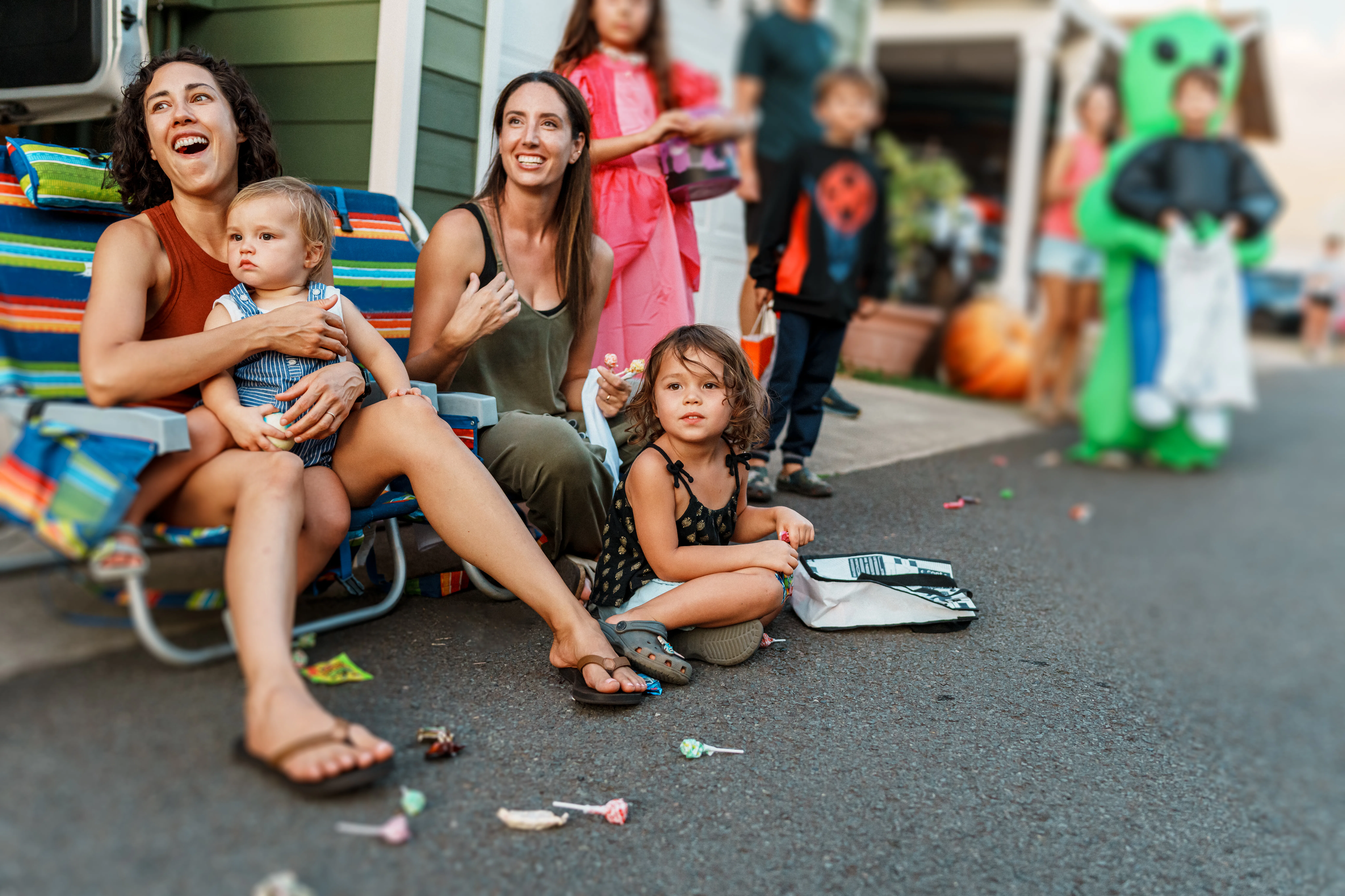 Two women and three young children seated on a curb during a neighborhood event, with costumed child...